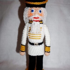 May include: A crocheted nutcracker doll with a white uniform, black pants, and a black hat with gold trim. The doll has a white beard and mustache, blue eyes, and a red mouth.