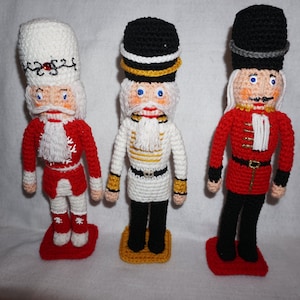 May include: Three crocheted nutcracker figurines. The nutcracker on the left is wearing a white hat with red and green trim, a red jacket, and white pants. The nutcracker in the middle is wearing a black hat with a gold band, a white jacket with gold trim, and black pants. The nutcracker on the right is wearing a black hat with a gray band, a red jacket with gold trim, and black pants.