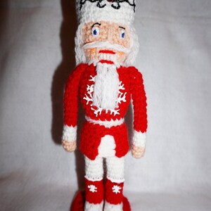 May include: A crocheted nutcracker doll with a white hat, red jacket, and white pants. The doll is standing on a red base.