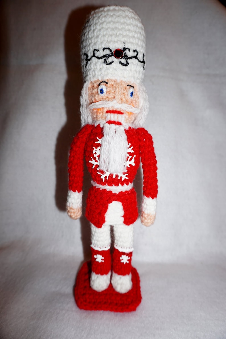 May include: A crocheted nutcracker doll with a white hat, red jacket, and white pants. The doll is standing on a red base.