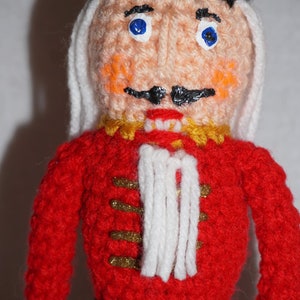 May include: A crocheted nutcracker doll with a red and gold uniform, white beard, and blue eyes. The doll is wearing a black hat with a gray trim.
