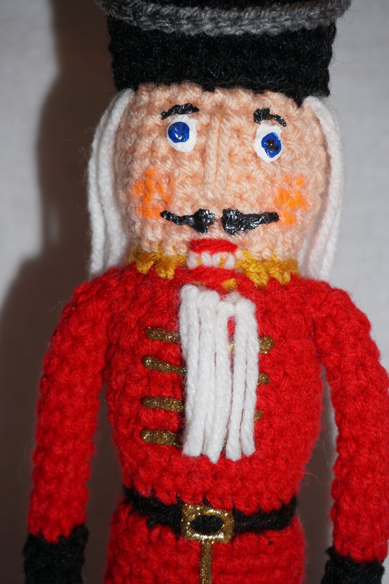 May include: A crocheted nutcracker doll with a red and gold uniform, white beard, and blue eyes. The doll is wearing a black hat with a gray trim.