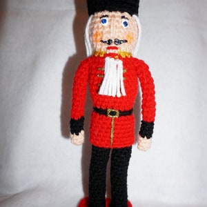 May include: A crocheted toy soldier with a red uniform, black hat, and white facial details. The soldier is standing on a red base.
