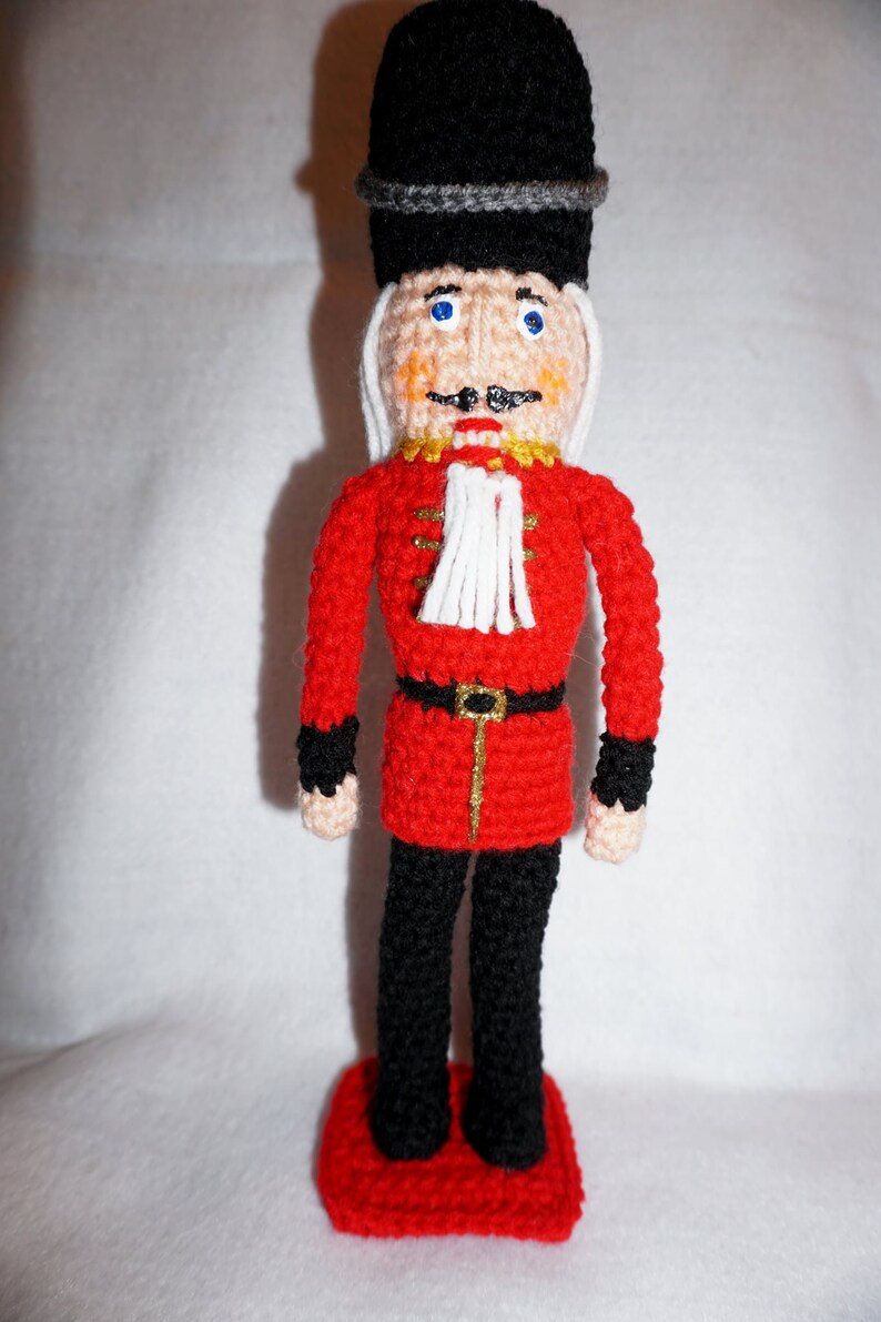 May include: A crocheted toy soldier with a red uniform, black hat, and white facial details. The soldier is standing on a red base.