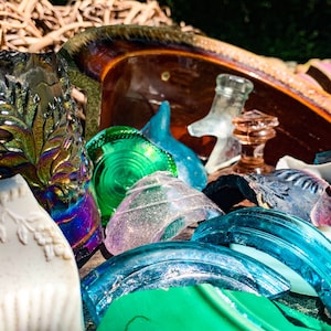 May include: A collection of broken glass and pottery pieces in various colors, including green, blue, white, and iridescent. The pieces are arranged in a pile on a brown surface.