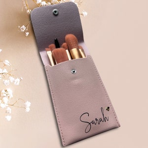 May include: A pink leather makeup brush holder with a snap closure and the name "Sarah" printed on the front. The holder is open and contains a set of six makeup brushes.