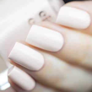 May include: Close-up of a hand with polished nails. The nails are painted in a light, creamy off-white color. The polish has a glossy finish. The background is blurred, with a glimpse of a nail polish bottle.