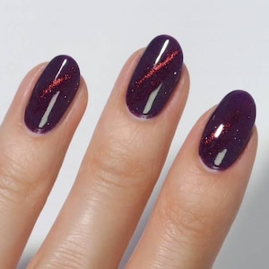 May include: Close-up of three fingernails painted with a deep purple nail polish. Each nail has a shimmering red stripe across the center. The nails are oval-shaped and have a glossy finish.