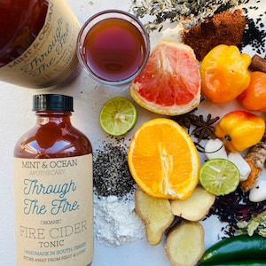 May include: An 8oz bottle of "Through The Fire" organic fire cider tonic with a black cap, next to a glass of the tonic. The image includes ingredients like citrus, ginger, peppers, and spices. The label reads "Handmade in South Jersey".