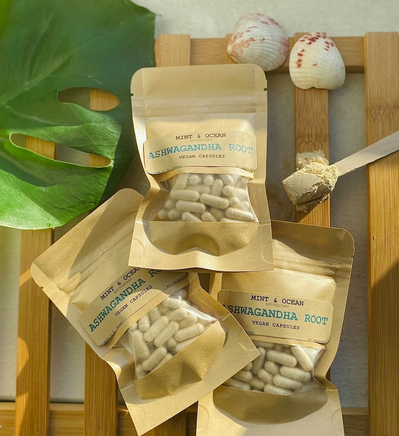 Op de afbeelding: Drie bruine papieren zakjes met doorzichtige plastic vensters die witte capsules bevatten. De zakjes zijn gelabeld met "Mint & Ocean Apothecary Ashwagandha Root Vegan Capsules".
