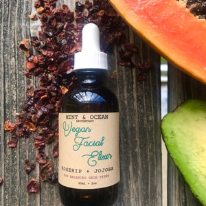 May include: A dark brown glass bottle with a white dropper top and a brown paper label. The label reads "MINT & OCEAN APOTHECARY Vegan Facial Elixir ROSEHIP + JOJOBA FOR BALANCED SKIN TYPES 60ml 2oz".