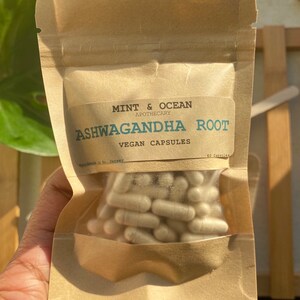 Op de afbeelding: Een bruine papieren zak met een wit label waarop staat "MINT & OCEAN APOTHECARY ASHWAGANDHA ROOT VEGAN CAPSULES 60 Capsules". De zak bevat witte capsules.