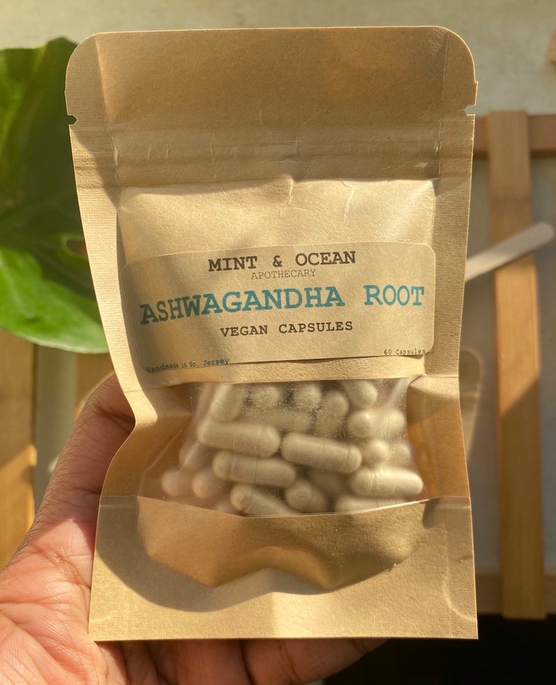 Op de afbeelding: Een bruine papieren zak met een wit label waarop staat "MINT & OCEAN APOTHECARY ASHWAGANDHA ROOT VEGAN CAPSULES 60 Capsules". De zak bevat witte capsules.