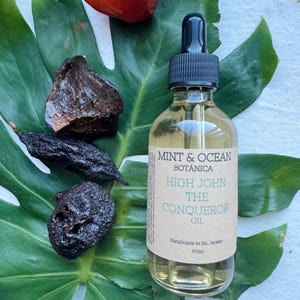 May include: A clear glass bottle with a black dropper top and a brown label that reads "MINT & OCEAN BOTÁNICA HIGH JOHN THE CONQUEROR OIL Handmade in So. Jersey 60ml". The bottle is sitting on a green leaf with a red, orange, and brown stone.