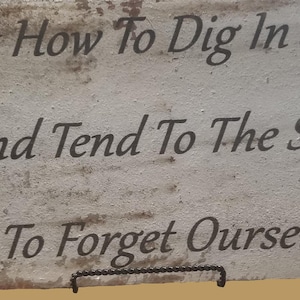May include: A rectangular, weathered sign with the text "To Forget How To Dig In The Earth, And Tend To The Soil, Is To Forget Ourselves" in dark brown lettering. The sign is supported by a black metal stand.