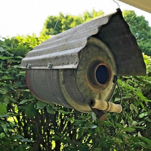 May include: A repurposed metal can birdhouse with a corrugated metal roof. The birdhouse is painted in shades of gray and brown and has a small round entrance hole. The birdhouse is hanging from a wire.
