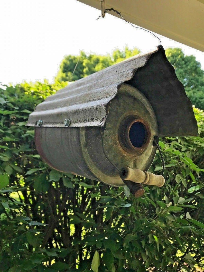 May include: A repurposed metal can birdhouse with a corrugated metal roof. The birdhouse is painted in shades of gray and brown and has a small round entrance hole. The birdhouse is hanging from a wire.