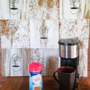 May include: A black coffee maker with a silver top and a red mug sitting on a wooden surface. A container of Coffee Mate French Vanilla creamer is next to the mug. The background is a wall with six square tiles that have a rustic, whitewashed look with the word "coffee" written on each tile.