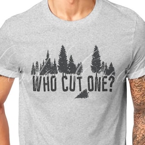 May include: Light gray t-shirt with a distressed black graphic of a forest and the text "WHO CUT ONE?". The shirt is made of a soft material and has short sleeves. A watch is visible on the left wrist.