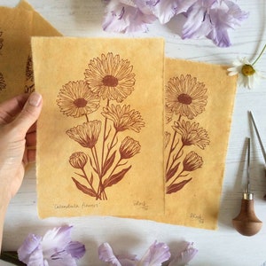 以下が含まれることがあります： 黄色い背景に、カレンデュラの花の植物画が2枚印刷されています。印刷物は「Calendula Flower」と「Black 1/50」というタイトルです。