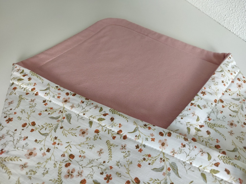 K&ouml;nnte beinhalten: Eine rosa-wei&szlig;e Wickelunterlage mit Blumenmuster. Die Unterlage ist in der H&auml;lfte gefaltet, so dass die rosa Seite oben und die Blumenmusterseite unten sichtbar ist.
