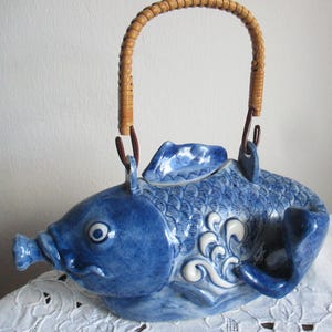 Könnte beinhalten: Eine blaue Keramik-Teekanne in Fischform mit detaillierter Schuppenstruktur, einem hervorstehenden Maul und einem Deckel in Form einer Rückenflosse. Die Teekanne hat einen geschwungenen, hellbraunen Rattangriff, der an zwei Metallösen befestigt ist.