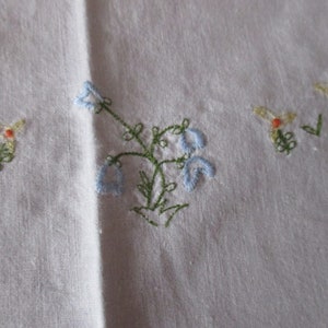 Puede incluir: Mantel de mesa de lino blanco con flores bordadas en azul y verde. Las flores est&aacute;n en un racimo y tienen un aspecto delicado y vintage.