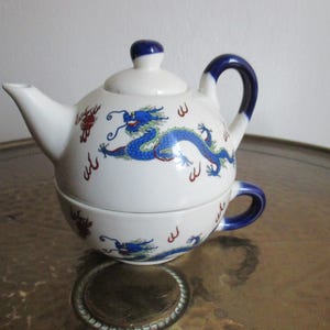 Könnte beinhalten: Ein weißes Keramik-Teeservice für eine Person, bestehend aus einer stapelbaren Teekanne und Teetasse. Das Set ist mit einem blau-grünen Drachenmotiv, roten Akzenten sowie einem dunkelblauen Henkel und Deckelknauf verziert.