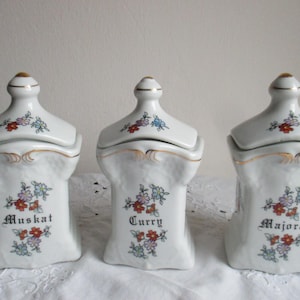 Três potes de porcelana antigos para especiarias, com os dizeres Manjerona, Noz-moscada e Curry em escrita gótica.