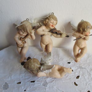 Puede incluir: Cuatro pequeñas figuritas de ángeles tocando instrumentos musicales. Los ángeles están hechos de porcelana blanca y tienen alas delicadas. Están sosteniendo un violín, una guitarra, una flauta y una trompeta. Los ángeles llevan todos túnicas blancas y tienen halos dorados.