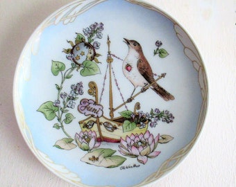 May include: A decorative porcelain plate with a blue background and gold trim. The plate features a hand-painted design of a bird perched on a small boat with flowers and leaves. The text "Faini" is written on the boat.