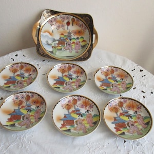 Könnte beinhalten: Ein Vintage-japanisches Porzellan-Teeservice mit einem Serviertablett und sechs kleinen Tellern. Jedes Stück hat einen Goldrand und zeigt eine farbenfrohe Szene mit Figuren, Gebäuden und Kirschblüten. Das Set wird auf einer weißen Spitzendecke präsentiert.