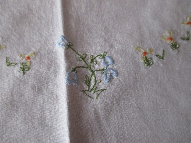 Puede incluir: Mantel de mesa de lino blanco con flores bordadas en azul y verde. Las flores est&aacute;n en un racimo y tienen un aspecto delicado y vintage.