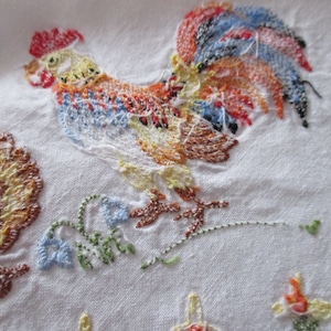 Puede incluir: Tela blanca bordada con un gallo colorido y una gallina marr&oacute;n. El gallo tiene una cresta y una barba rojas, una cola azul, negra y naranja, y un cuerpo amarillo. La gallina es marr&oacute;n y amarilla. La tela est&aacute; decorada con hojas verdes y flores azules.