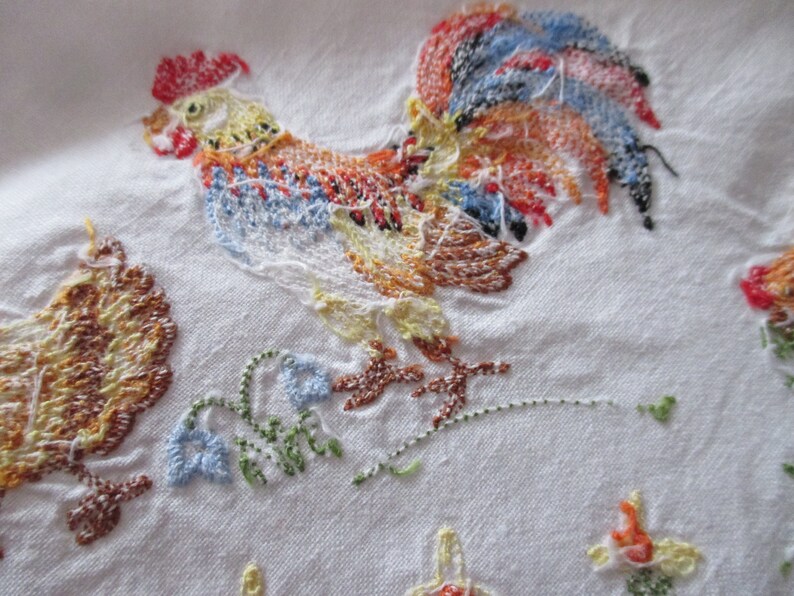 Puede incluir: Tela blanca bordada con un gallo colorido y una gallina marr&oacute;n. El gallo tiene una cresta y una barba rojas, una cola azul, negra y naranja, y un cuerpo amarillo. La gallina es marr&oacute;n y amarilla. La tela est&aacute; decorada con hojas verdes y flores azules.