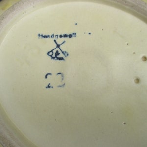 Puede incluir: Primer plano de la parte inferior de un plato de cer&aacute;mica con un dise&ntilde;o pintado a mano en azul y blanco. El plato tiene un esmalte amarillo y una marca azul que dice "Handgemacht" con un s&iacute;mbolo de molino de viento y el n&uacute;mero "22".