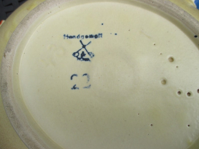 Puede incluir: Primer plano de la parte inferior de un plato de cer&aacute;mica con un dise&ntilde;o pintado a mano en azul y blanco. El plato tiene un esmalte amarillo y una marca azul que dice "Handgemacht" con un s&iacute;mbolo de molino de viento y el n&uacute;mero "22".