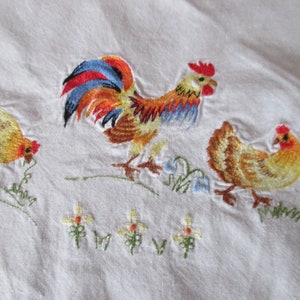 Puede incluir: Toalla de cocina de lino blanco bordada con un gallo y dos gallinas. El gallo es rojo, naranja, azul y marr&oacute;n. Las gallinas son marrones y amarillas.