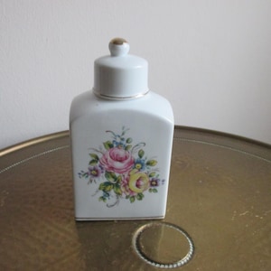 Könnte beinhalten: Weiße Porzellanflasche mit goldenem Deckel und Blumenmuster. Die Flasche zeigt einen Strauß aus rosa, gelben und weißen Blumen mit grünen Blättern.