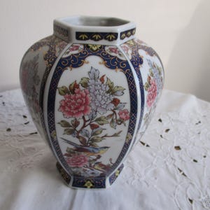 Puede incluir: Un jarrón de porcelana blanca con forma hexagonal y un diseño floral. El jarrón presenta flores azules, doradas y rosas con un pájaro posado en una rama.
