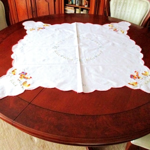 Puede incluir: Mantel blanco bordado con un borde festoneado y un dise&ntilde;o de gallos y flores. El mantel est&aacute; colocado sobre una mesa redonda de madera.