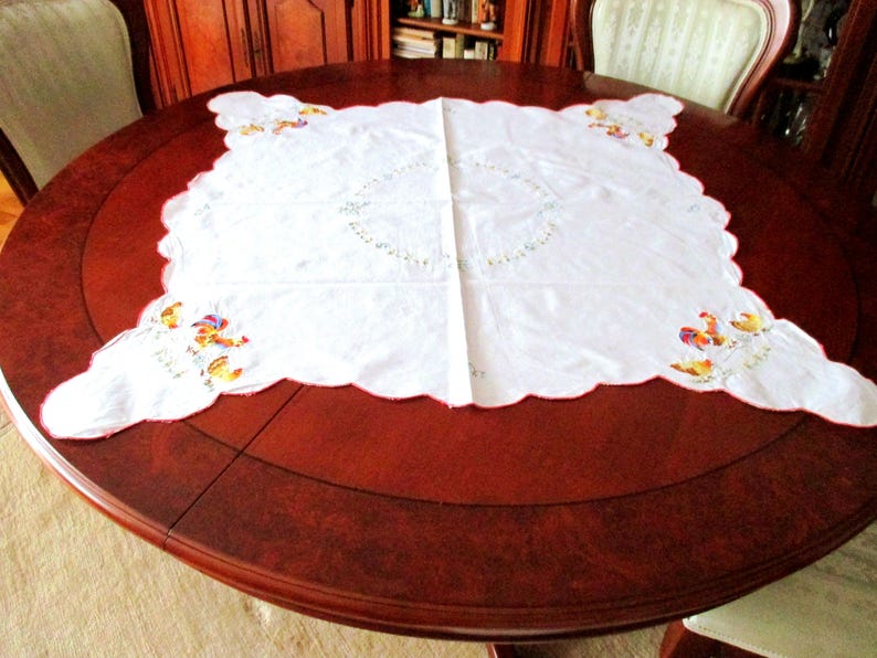 Puede incluir: Mantel blanco bordado con un borde festoneado y un dise&ntilde;o de gallos y flores. El mantel est&aacute; colocado sobre una mesa redonda de madera.