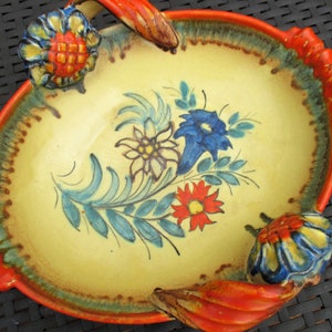 Puede incluir: Un plato de servir de cer&aacute;mica amarillo con dos asas naranjas y dise&ntilde;os florales en azul, rojo y blanco. El plato tiene un aspecto r&uacute;stico y pintado a mano.
