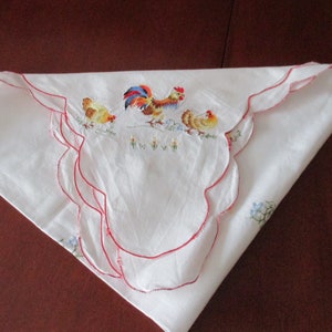 Puede incluir: Mantel blanco con un borde festoneado rojo y un dise&ntilde;o bordado de un gallo y dos pollitos.