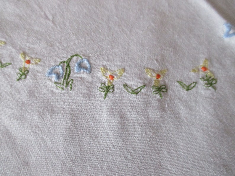 Puede incluir: Tela blanca con flores bordadas en tonos azules, amarillos y naranjas. Las flores est&aacute;n dispuestas en una fila a lo largo del borde inferior de la tela.