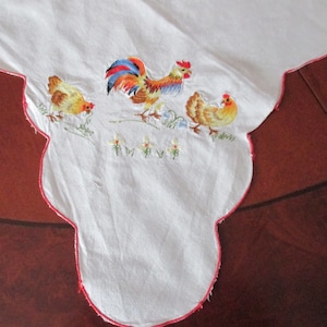 Puede incluir: Mantel blanco con un borde festoneado rojo y bordado con un gallo y dos gallinas. El gallo es rojo, amarillo y azul. Las gallinas son amarillas.
