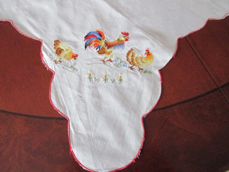 Puede incluir: Mantel blanco con un borde festoneado rojo y bordado con un gallo y dos gallinas. El gallo es rojo, amarillo y azul. Las gallinas son amarillas.