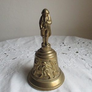 Könnte beinhalten: Eine Vintage-Messingglocke mit einer Figur oben. Die Glocke hat ein detailliertes Design von Menschen, die einen Sport um den Sockel spielen. Die Figur oben trägt einen Hut und einen Mantel. Die Glocke ist wahrscheinlich aus Messing.