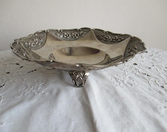 May include: Silver-toned, ornate serving dish with a scalloped edge and floral detailing. The dish has a shallow bowl and rests on a decorative pedestal base. The surface reflects light, highlighting its polished finish. A vintage piece for serving or display.
