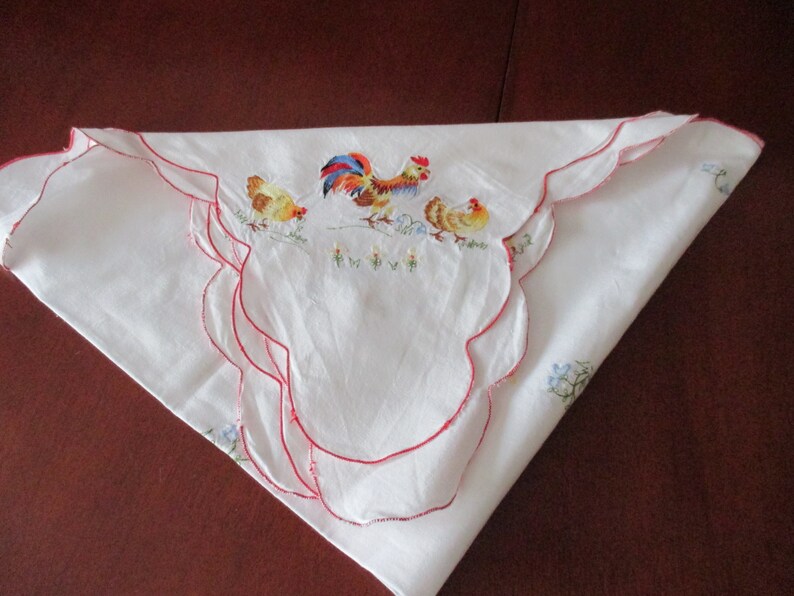 Puede incluir: Mantel blanco con un borde festoneado rojo y bordado con un gallo y dos pollitos. El gallo es rojo, azul y amarillo, y los pollitos son amarillos.